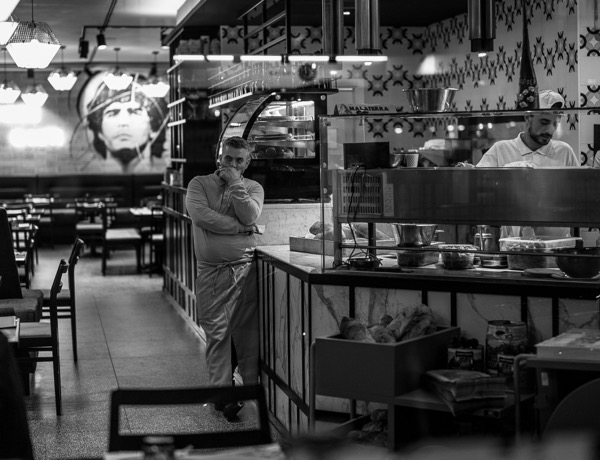 A restaurant owner leaning against the counter contemplating as as staff member works behind the counter.