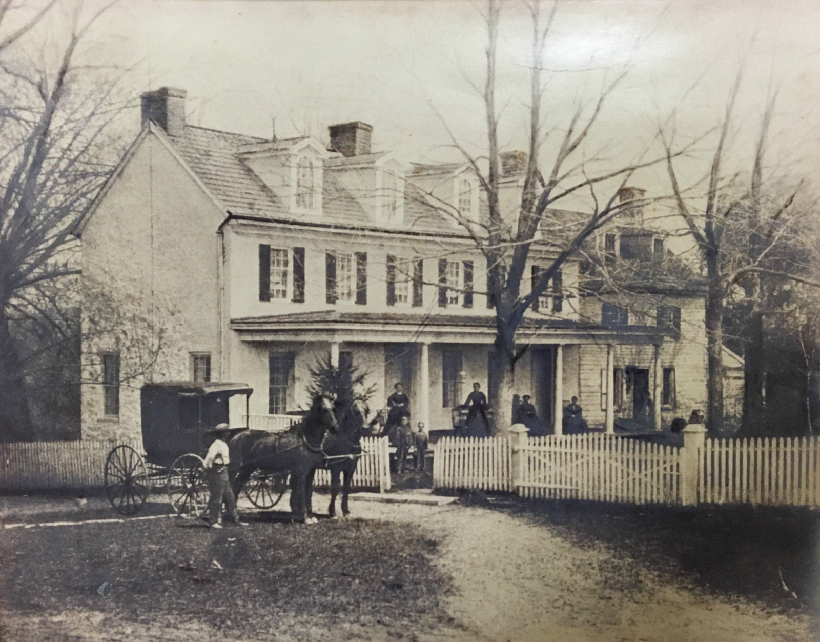 A black and white photo of the front of a British cottage with people and horses.