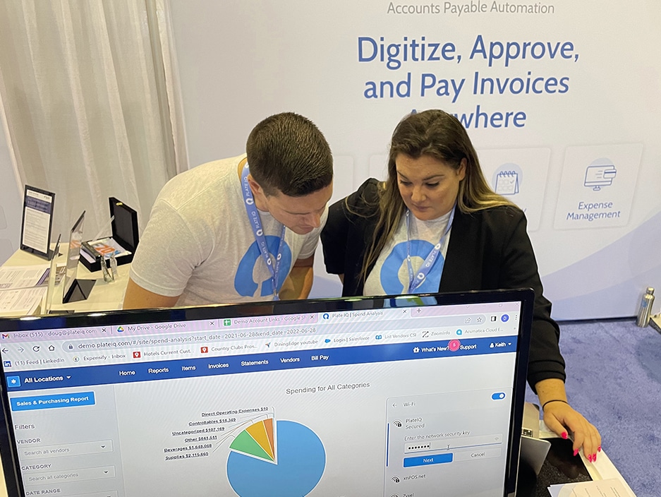 Colleagues adjust a demo screen at Ottimate's booth in Orlando, Florida, at the HITEC hospitality conference for hotels and country clubs. Background explains that Ottimate's accounts payable automation means that hotel groups and country clubs can digitize, approve and pay invoices from anywhere. 