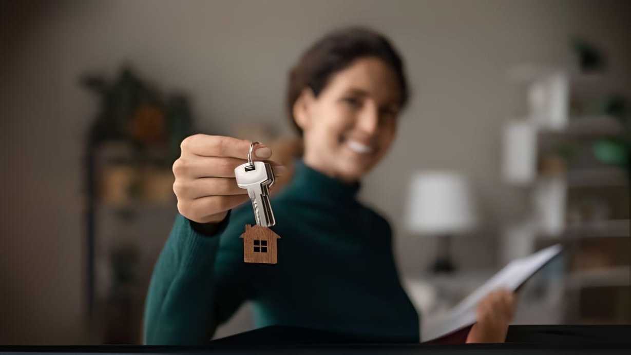 Smiling real estate woman holding up keys with a wooden house in focus hanging from the key ring